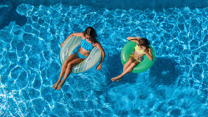 Aerial top view of children in swimming pool from above, happy kids swim on inflatable ring donuts and have fun in water on family vacation
