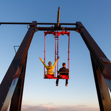Young Couple On The Swing On The Roof Of Tower At Sunset Time