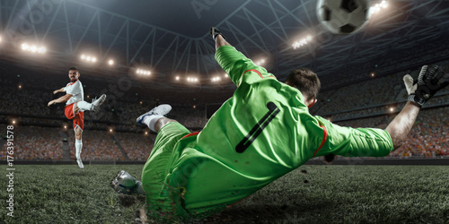 Zdjęcie XXL Piłkarz uderza piłkę do bramki na profesjonalnym stadionie. Bramkarz chroni bramę do piłki nożnej. Gracze mają na sobie niezarejestrowany mundur sportowy.