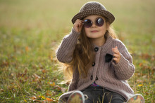 Little Girl With Sunglasses And Hat Sitting On A Grass In Autumn Clothes