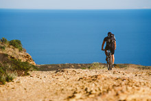 A Young Guy Riding A Mountain Bike On A Bicycle Route In Spain On Road Against The Background Of The Mediterranean Sea. Dressed In A Helmet, A Dark One And A Black Backpack