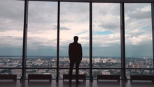 Rear View Of Man In Formal Suites Who Stand In Front Of Panoramic Window With City View. A Man Stands In Front Of Big Panoramic Wiindow View From Back.