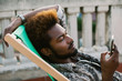 © BONNINSTUDIO/Stocksy - Young african american man using his smartphone sitting on a deck chair on a rooftop.