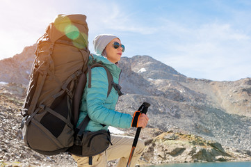 A hiker girl in sunglasses and a hat with a backpack and mountain gear with tracking treks in her hands looks at the beautiful view of a high mountain lake in an archipelago in the northern Caucasus