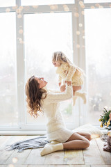 A beautiful mother is playing with happy daughter at the panoramic window in the knitted sweater near the Christmas tree. A young woman hugs little girl, smiling. New year festive card. copy space