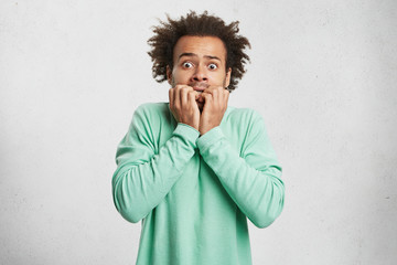 Wall Mural - Portrait of young Afro American man with scared and anxious expression, bites finger nails, wears green sweater, being afraid of visiting doctor. Human facial expressions and negative emotions concept