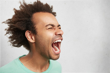 Horizontal portrait of man with dark skin and Afro hairstyle screams in despair, opens mouth widely, being in panic. Frustrated mixed race man poses against white studio background with copy space