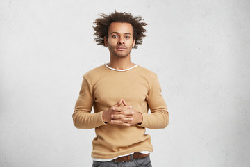 Wall Mural - Indoor shot of confident mixed race businessman dressed casually, keeps hands together, looks convincely, going to present project to partners. Stylish man with curly hair, poses in white studio