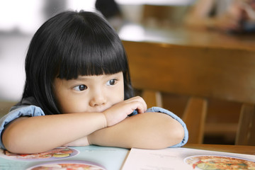 Asian children cute or kid girl hungry and waiting order or food for breakfast or lunch on menu and wood table with chair at cafe restaurant with space