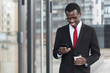 © Damir Khabirov - Indoor portrait of busy African chief executive officer pictured in afternoon, holding smartphone in hands and coffee cup, looking attentively at screen with satisfied happy smile while checking web