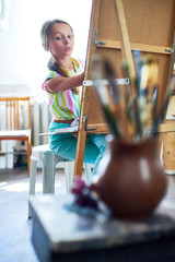 Wall Mural - Portrait of talented little girl painting still life picture on easel in art class looking at art supplies set up