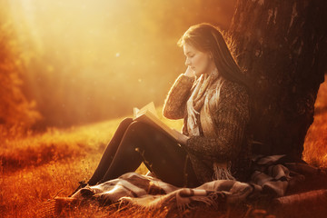 Wall Mural - Beautiful young brunette sitting on a fallen autumn leaves in a park, reading a book