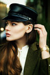 Close up outdoor portrait of young beautiful woman with long hair wearing trendy military leather cap, wrist watch, earrings. Model looking aside, posing in street. Female fashion concept
