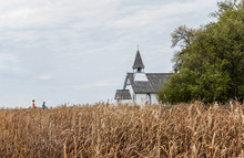 Horizontal Distant Full Length Image Of Two Caucasian Cowboys Walking Toward A Beautiful Little Country Church Half Hidden By A Tall Harvest Ready Golden Field.