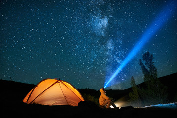 man tourist with flashlight near his camp tent at night.