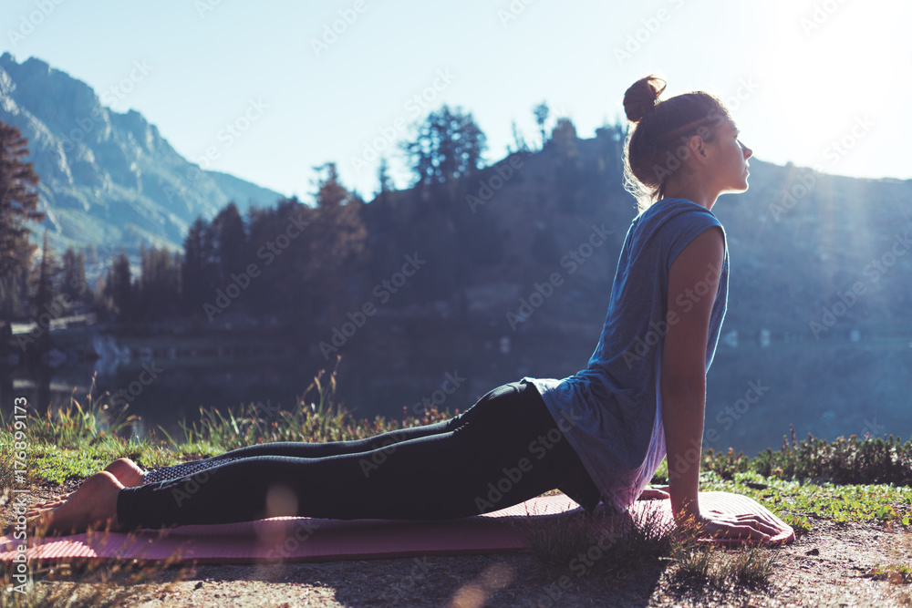Fit young happy traveling girl practicing yoga outdoor in the stunning mountain wilderness in front of amazing cold lake at sunrise. Calmness and relax, female happiness. - obrazy, fototapety, plakaty 