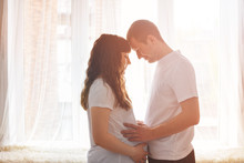 Happy Young Couple Expecting Baby Standing Together In Front Of Window Holding A Belly And Thinking Of A Baby