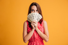 Young Thinking Pretty Woman In Red Dress Hiding Behind Bunch Of Money Banknotes, Looking Upward
