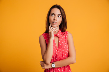 Wall Mural - Photo of thinking young woman in red dress touching her chin, looking upward, isolated on yellow background