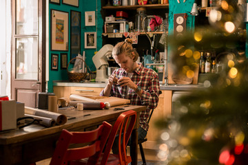 Young adult man with long blonde hair wrapping christmas present with red ribbon seated on wooden table in cozy apartment indoor.