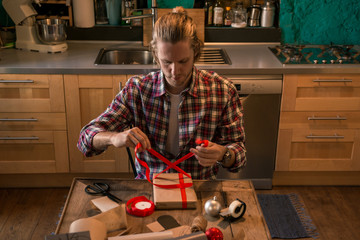 Young adult man with long blonde hair wrapping christmas present with red ribbon seated on wooden table in cozy apartment indoor.