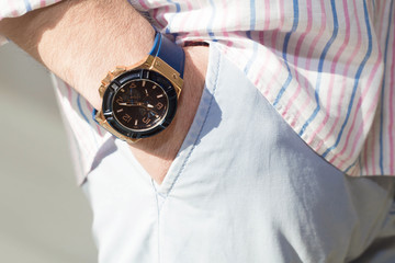  fashion image of luxury watch on wrist of man.body detail of casual look.Man's hand in blue pants pocket closeup at white background.Man wearing striped blue pink shirt and blue pants.Not isolated