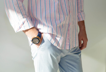 Wall Mural - fashion image of luxury watch on wrist of man.body detail of casual look.Man's hand in blue pants pocket closeup at white background.Man wearing striped blue pink shirt and blue pants.Not isolated