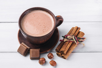 Hot cocoa with milk in brown clay cup, broken chocolate cubes, hazelnut and cinnamon sticks on white wooden planks