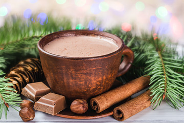 Blurred Christmas lights, cup of hot cocoa with milk, broken chocolate cubes, hazelnut and cinnamon sticks, christmas tree branches, pine cone on white wooden planks