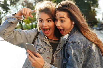 Poster - Close-up portrait of two surprised young pretty brunette woman looking at mobile phone, outdoor