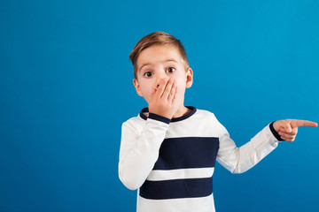 Surprised young boy in sweater pointing away and covering mouth