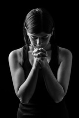 Wall Mural - Faithful athletic woman praying, with hands folded in worship to god, head down and eyes closed in religious fervor, on low key black background. Concept for religion, faith, prayer and spirituality
