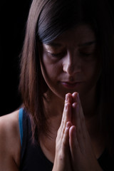 Wall Mural - Faithful athletic woman praying, with hands folded in worship to god, head down and eyes closed in religious fervor, on black background. Concept for religion, faith, prayer and spirituality
