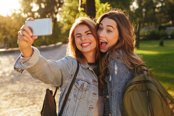 Wall Mural - Close-up photo of two funny girls in casual jeans wear making selfie on smartphone, outdoor