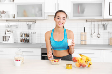 Wall Mural - Young woman in sportswear drinking milk and eating oatmeal in kitchen