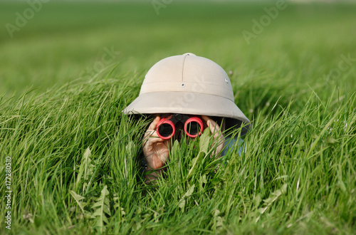 Fotografia Little Boy with Binoculars and safari Hat, laying in the grass searching for Kno