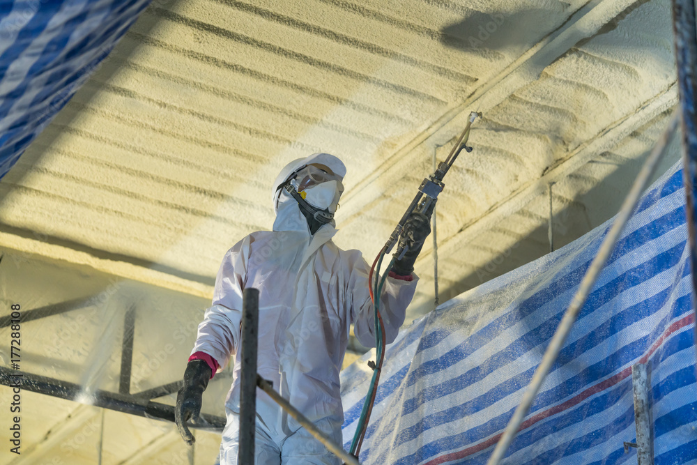 Technician spraying foam insulation using Plural Component Gun for polyurethane foam - Repair tool in the white protect suit applies a construction foam from the gun to the roof of a warehouse. - obrazy, fototapety, plakaty 