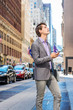 © Alexander Image - Young Businessman traveling, working in New York, wearing gray blazer, patterned shirt, carrying laptop computer, holding cell phone, standing on street in Manhattan, looking up, thinking..