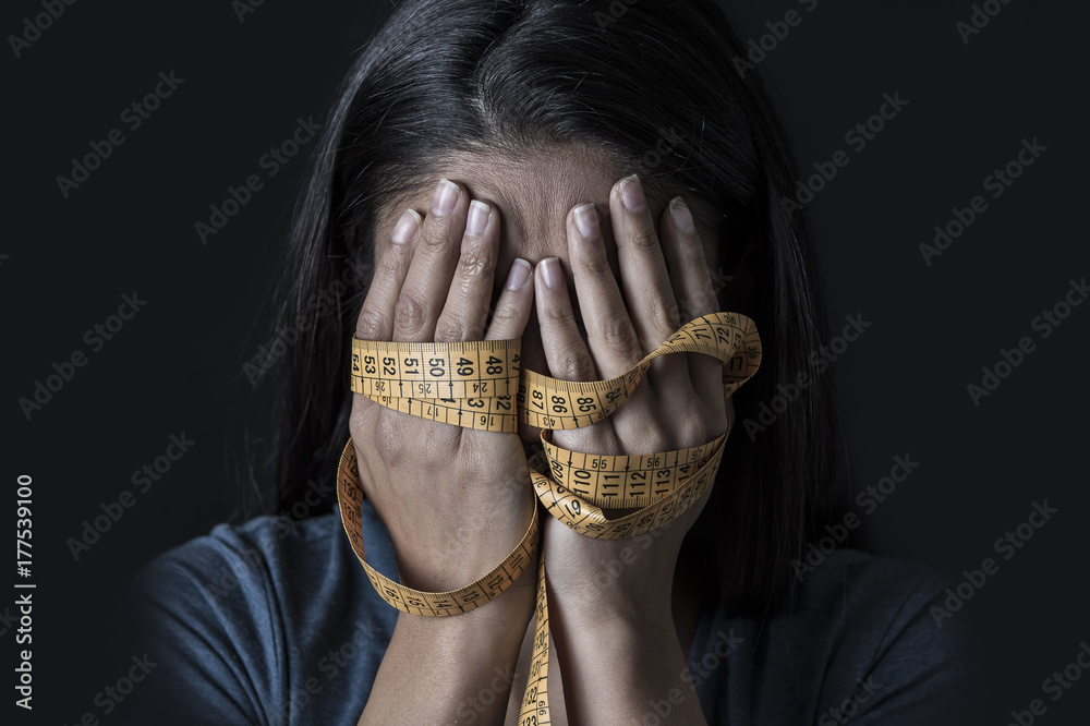 hands wrapped in tailor measure tape covering face of young depressed and worried girl suffering anorexia or bulimia nutrition disorder - obrazy, fototapety, plakaty 