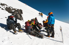 Fully Equipped Professional Mountaineers On A Stop Sit On A Snowy Slope In Sunny Weather.