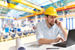 © industrieblick - Ingenieur im Maschinenbau telefoniert am Arbeitsplatz/ Schreibtisch in einer Fabrikhalle und arbeitet am Rechner