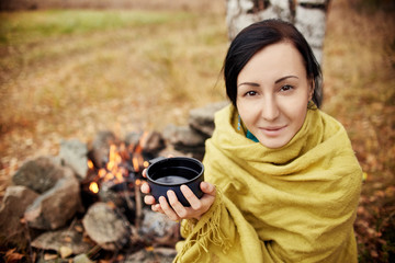 Portrait of a woman with a mug of hot tea in his hands autumn in a forest campfire. A picnic in the autumn forest. Girl wrapped in a blanket warmed in a forest bonfire