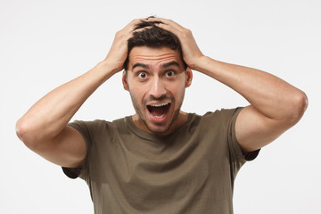 Headshot of handsome surprised manlooking at the camera, astonished with big sale prices, holding hands on head, standing against grey background. Human face expression concept