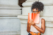 Young African American Female College Student With Afro Hairstyle, Wearing Glasses, Standing On Street By Vintage Style Column In New York, Holding Red Book, Covering Face, Looking Away, Thinking..