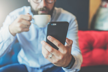 Wall Mural - Close-up of black smartphone in man's hand. In background, in soft focus, young bearded businessman drinks coffee. Guy uses digital gadget, checking email, reading, blogging. Online working,education.