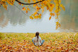 © Iryna - Back view of young woman with two braids sitting at foliage and looking at water on shore of a lake or river. Branch with yellow leaves above her.