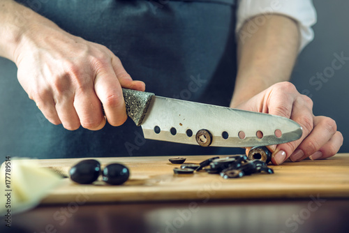 Zdjęcie XXL Szef kuchni w czarnym fartuchu przecina czarne oliwki nożem. Koncepcja ekologicznych produktów do gotowania