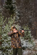 © scharfsinn86 - Male hunter in camouflage and with backpack, armed with a rifle, standing in a snowy winter forest.