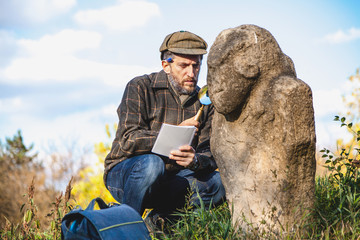 Scientific historian examines through magnifying glass stone sculpture on mound