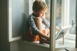 © Iryna - Sad rose cheeks barefoot smiling preschooler boy dressed home clothes sitting on windowsill and looking on foto in the frame.